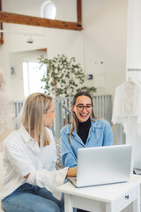 Two women sitting on a couch, working on a laptop in a bright, modern space with white curtains and clothing racks in the background.