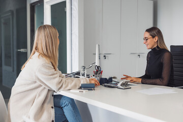 Fototapeta premium A young woman with long blonde hair sits at a desk, smiling while looking at a computer screen. She is wearing a cozy beige jacket and has a thoughtful expression. The workspace is modern and bright,