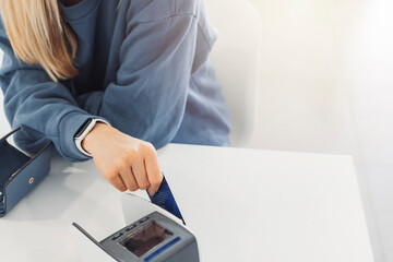 A woman with long blonde hair is smiling while using a card reader at a table. She is wearing a blue sweatshirt and appears to be in a bright, modern space. A wallet is visible on the table.