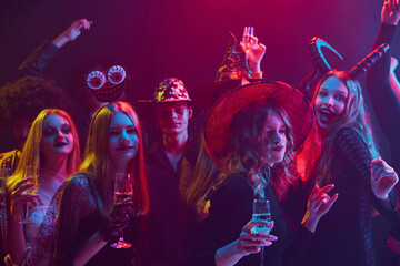 Lively group of friends, men and women attending Halloween party, dressed in various costumes of supernatural character, laughing and dancing under colorful lights in nightclub. Concept of Halloween