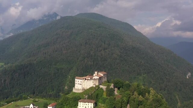 Slow flight to the Castle of Stenico, with mountains in the clouds and green trees in the background. Panoramic view of Stenico Castle in the Valli Giudicarie, Trentino, with lush green surroundings.