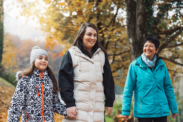 Three women walking hand in hand through a park covered in autumn leaves. They are smiling and enjoying the fall scenery, with colorful trees in the background.