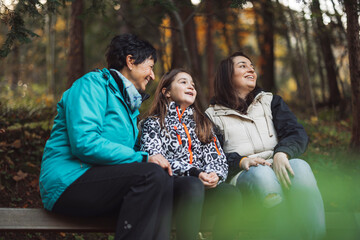 A joyful moment shared between three generations of women in a forest setting. The older woman smiles at her granddaughter, who is animatedly speaking, while another woman listens with a warm expressi