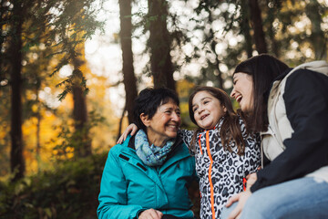 A joyful moment shared between three generations of women in a forest setting. The older woman smiles at her granddaughter, who is animatedly speaking, while another woman listens with a warm expressi