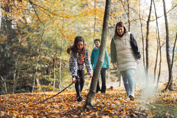 A young girl in a leopard print jacket explores a forest path, while two women walk behind her. The scene is set in autumn with colorful leaves on the ground and trees in the background.