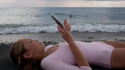 Beautiful teenage girl using smartphone while lying down on beach near the sea. Copy space - Powered by Adobe