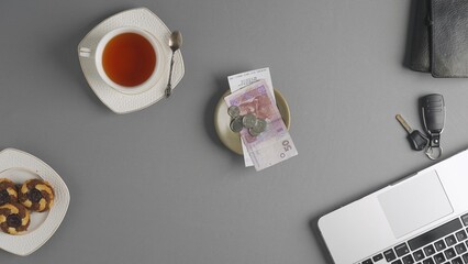 Top view of a saucer with a check for payment, coins and hryvnia bills, next to a cup of tea and a laptop, wallet and car keys on the table. The concept of money and payment for services.