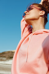 joyful woman in pink sweatshirt and sunglasses raises her arms in celebration on sandy beach