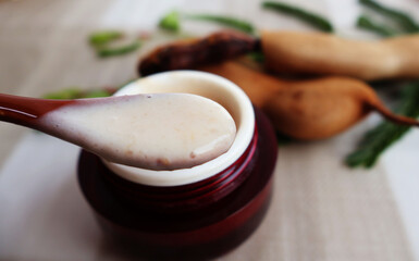 Use a spoon to scoop out the brown cream. from the red jar Next are ripe tamarind leaves and pods on a light-colored table.

