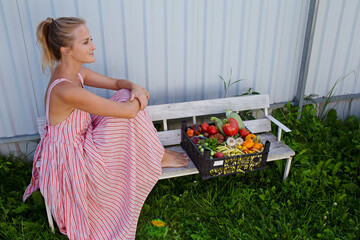 A beautiful girl with a basket of fresh vegetables sits on a white bench. This is a moment of rest after harvesting, which fills the girl with joy and pride in her work