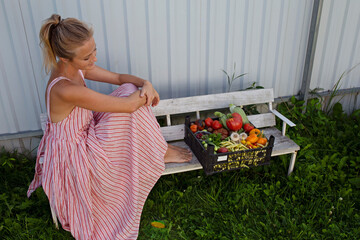 A beautiful European girl is sitting on a white bench, and next to her is a basket of fresh vegetables