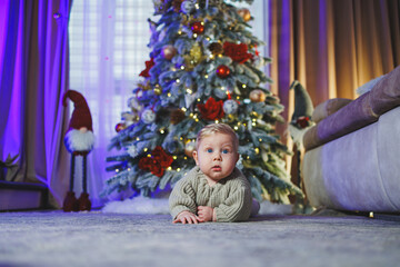 A baby in a beige knitted sweater on the background of a Christmas tree. Baby's first Christmas. A small child on the background of Christmas decorations.