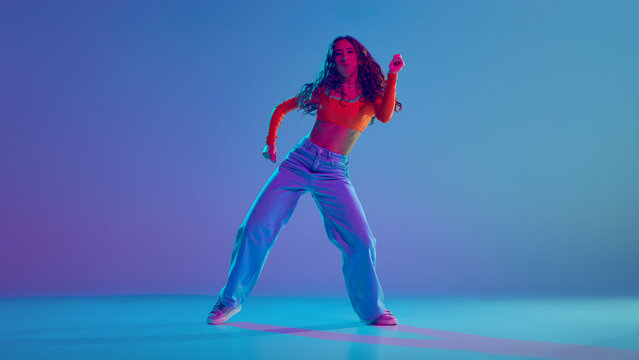 Young girl, dancer energetically dancing hip hop during her dance training against gradient blue background in neon light. Concept of freestyle dance, modernity, urban culture, youth