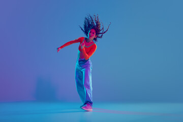 Hip hop training session in full motion, female dancer performing with passion against gradient blue background in neon light. Concept of freestyle dance, modernity, urban culture, youth