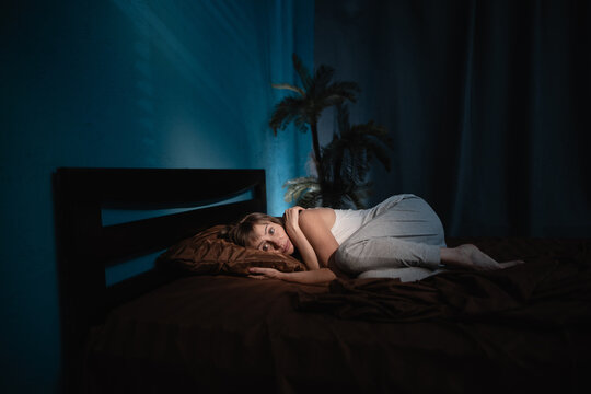 An elderly woman who woke up in the middle of the night from a nightmare or panic attack lies curled up on her bed in her bedroom, frightened