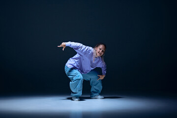 Artistic and talented young girl, dancer in motion against navy blue studio background. High-energy hip hop performance. Concept of freestyle dance, modernity, urban culture, youth