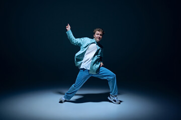 Stylish young guy, hip-hop dancer in motion on stage, training, performing with energy against navy blue background. Concept of freestyle dance, modernity, urban culture, youth