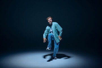 Dynamic image for young man in casual clothes in motion, dancing hip-hop against navy blue background. Shadows. Concept of freestyle dance, modernity, urban culture, youth