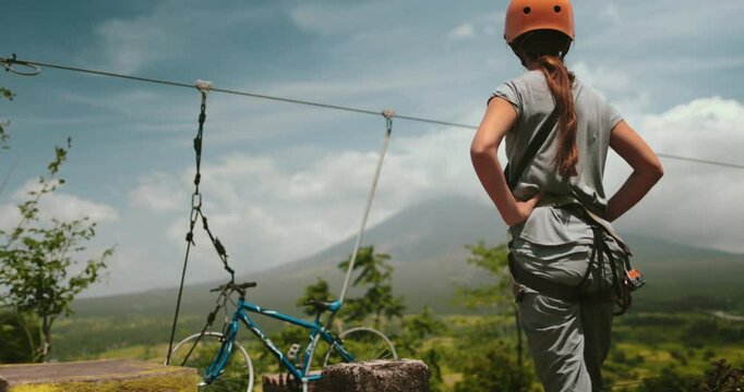 Young woman wearing a helmet is standing next to a zip line with a volcano in the background. She is about to embark on an exhilarating ride through the air