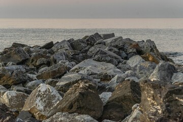 A rocky shoreline with large boulders extending into the calm sea under a hazy sky