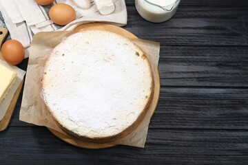 Delicious sponge cake with powdered sugar and ingredients on black wooden table, flat lay. Space for text