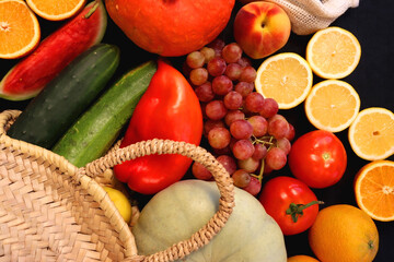 Round straw bag and various seasonal fruits and vegetables on dark background. Summer and fall produce. Top view.