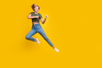Full size photo of active girl dressed khaki crop top jeans jump clenching fists look empty space isolated on yellow color background