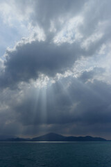 A serene moment unfolds as rays of sunlight break through dramatic clouds, illuminating the calm sea and distant mountains, creating a peaceful natural atmosphere