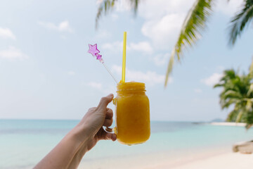 A sun-soaked beach scene shows a person enjoying a vibrant tropical drink while standing near the shoreline, surrounded by palm trees and gentle waves