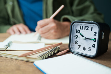A clock on the table with a man is writing on the notebook, writing and time concept