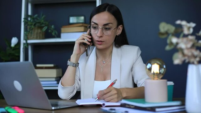 Professional businesswoman works at her laptop, talking on the phone while writing in a notebook. She is surrounded by office supplies, using a laptop, and sitting in a modern, stylish workspace.