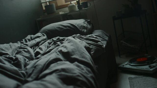Unmade bed with dark grey rumpled sheets and blankets, small bedside table near it, record player on floor