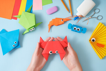 Female hands holding finished origami paper toy crab. On the table are other finished toys fish, stingray, turtle, shrimp. DIY project concept for kids. Flat lay