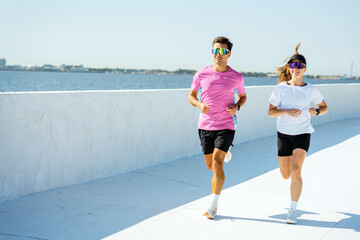 Fototapeta premium A fit couple runs energetically along a waterfront path, with the city skyline visible in the distance.