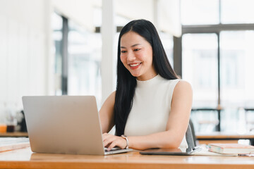 Asian woman working on laptop in bright office, smiling happily