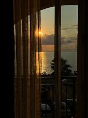 Sunset view through a window with sheer curtains overlooking the sea.