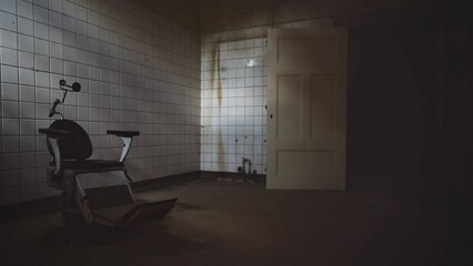 Dimly lit, abandoned room with white, tiled walls and a solitary dental chair in the center