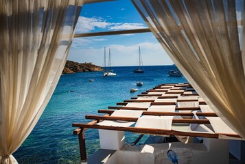 white curtains framing a sun-drenched terrace overlooking a turquoise sea with sailboats