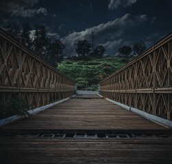 Long, wooden bridge with metal railings, leading towards a hillside shrouded in ominous clouds