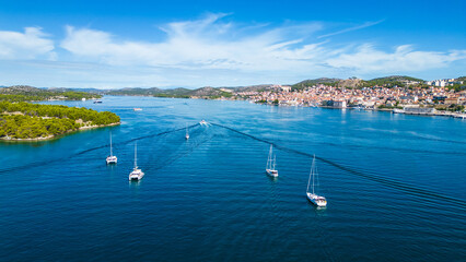 Obraz premium A yacht departs from the marina in Šibenik, Croatia, gliding through the calm waters of the Adriatic Sea. This aerial view captures the scenic coastline, with the historic city of Šibenik 