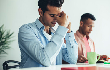 Fototapeta premium Concerned guy sitting in office rubbing nose in thoughts