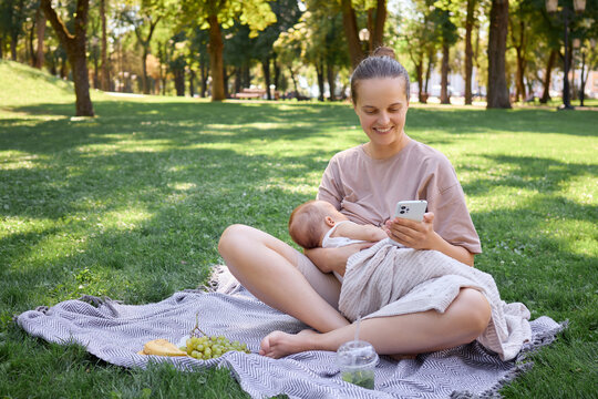 Loving mother breastfeeding her baby in park enjoying summer picnic on green grass and using smartphone for social media browsing and chatting or shopping online