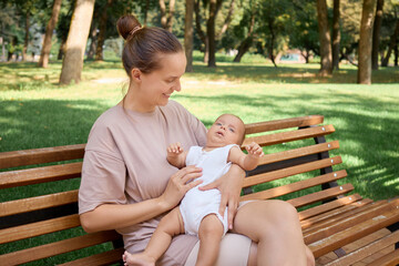 Attractive smiling woman with bun hairstyle spending joyful day in park her infant son or daughter sitting on bench in park in peaceful outdoor setting