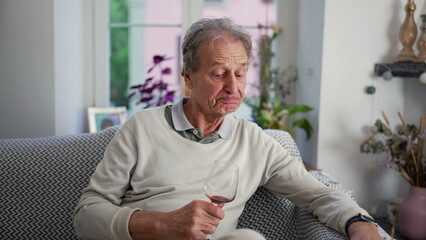 Elderly man taking a sip of red wine while sitting on a sofa, captured in a cozy home setting