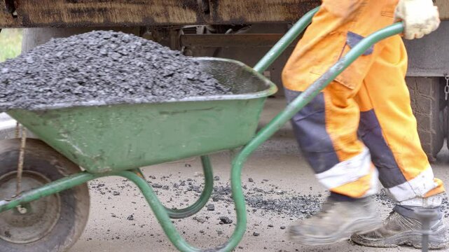 Road workers unload hot asphalt from delivery vehicle into wheelbarrow and shift it to place of work