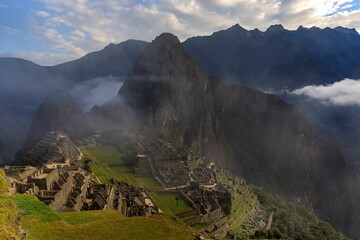 Machu Picchu with misty mountains and a partly cloudy sky.