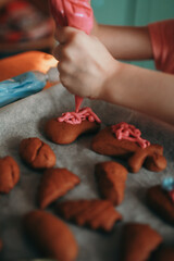 A child in a pink T-shirt is piping pink icing onto gingerbread cookies, with various other cookies resting on parchment paper nearby.