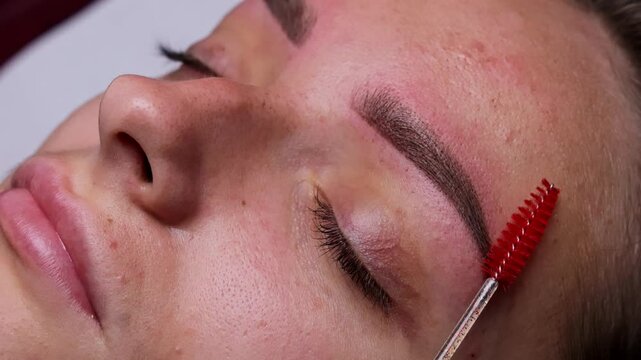Relaxed girl with closed eyes after permanent eyebrow makeup master brushes eyebrows with a brush Eyebrow tattoo procedure.