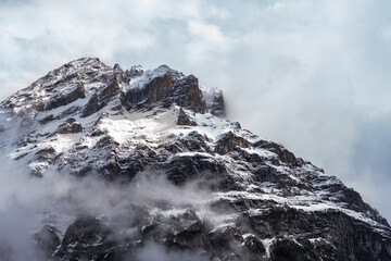 Landscape  of a snowy mountain peak among the Alps in Grindelwald, Switzerland. Snow covers the rocky face of the mountain, and the sky is blue above with soft white clouds.