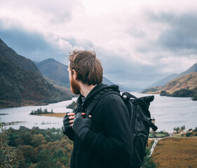 A male hiker looks out over the distant hills and autumn landscape near the Glenfinnan Monument in the Highlands, Scotland. He wears a backpack, hoodie, and fingerless gloves.